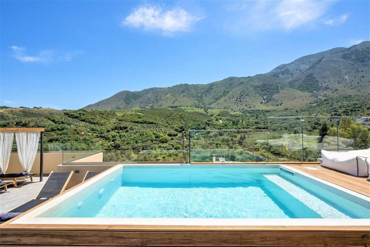 A rooftop infinity pool with a wooden deck, lounge chairs, and a view of green hills and mountains.