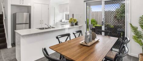 Cocina moderna con armarios blancos, un refrigerador de acero inoxidable y una zona de comedor con mesa de madera y sillas negras.