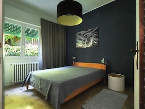 Un dormitorio con cama, un radiador, una ventana con cortinas, una lámpara de pared y un cuadro.