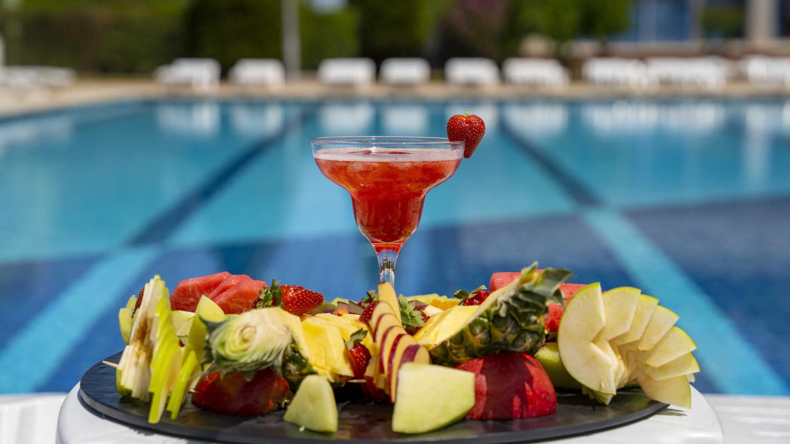 A fruit platter with a cocktail on a table by a pool.