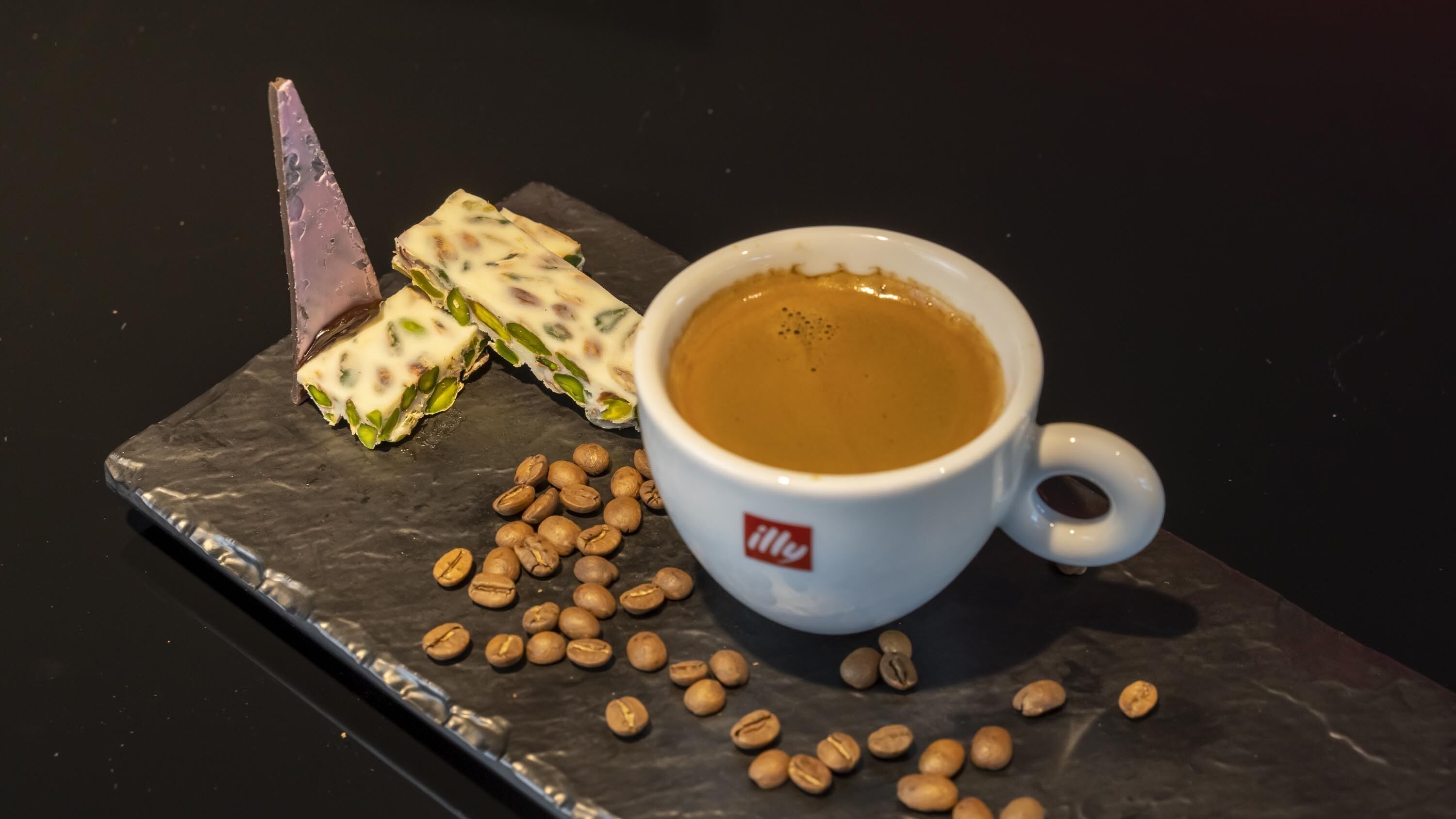 A cup of coffee with a logo, a chocolate dessert, and scattered coffee beans on a slate surface.