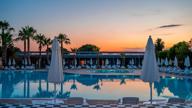 A pool area with lounge chairs and umbrellas, palm trees, and a sunset in the background. - Amon Hotel Belek - All Inclusive (Serik)