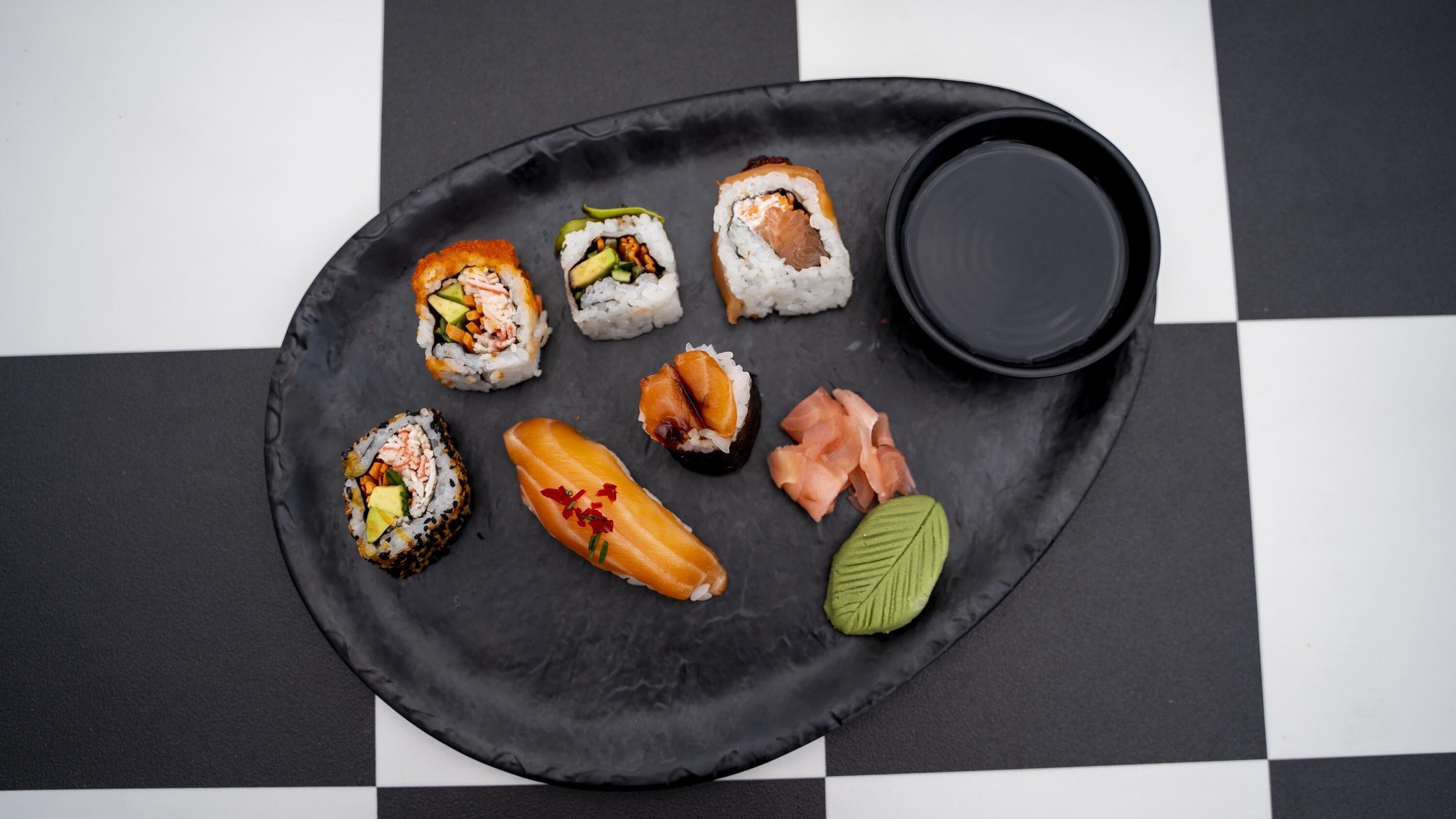 A black plate with various sushi pieces and a small bowl of sauce on a checkered tablecloth.