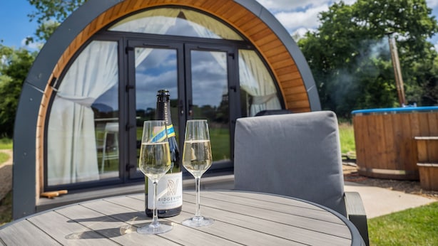 Une structure circulaire moderne en bois, dotée d’une porte et de fenêtres en verre, une bouteille de vin et deux verres posés sur une table en bois.