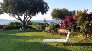 Een tuin met een witte lounge stoel, een grote boom en uitzicht op zee.