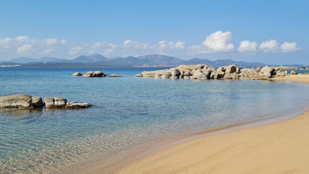 Een strand met helder water, grote rotsen en bergen op de achtergrond.