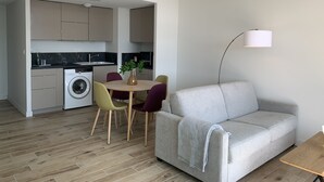 A modern living room with a grey sofa, a round wooden table with chairs, a washing machine, and a kitchen area with cabinets and a sink. - THE LAVOISIER TERRACE (La Rochelle)