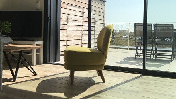 A modern living room with a yellow armchair, a TV on a wooden stand, and a large sliding glass door leading to an outdoor patio with a table and chairs. - THE LAVOISIER TERRACE (La Rochelle)