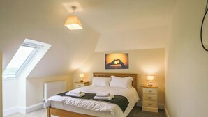 Un dormitorio con techo inclinado, tragaluz, cama con sábanas blancas, dos mesitas de noche con lámparas, y un cuadro en la pared.
