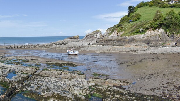 A rocky beach with a boat, cliffs, and a grassy hill.