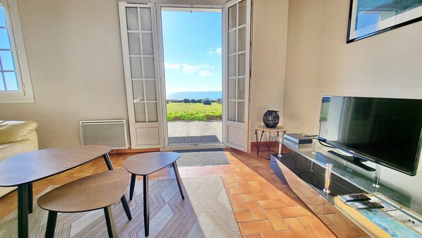 A living room with a TV, a sofa, a dining table, and chairs. There is a large window with a view of the outdoors.