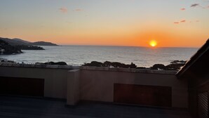 A sunset view from a balcony overlooking a coastal area with rocky terrain and distant hills.