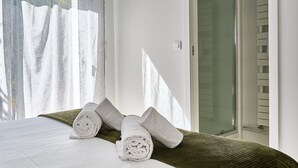 Una cama bien hecha con toallas blancas enrolladas, un edredón verde texturizado y un vidrio que da a la zona de la ducha.