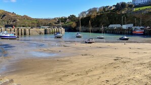 Escena costera con barcos fondeados cerca de una playa de arena, un puente y edificios al fondo.