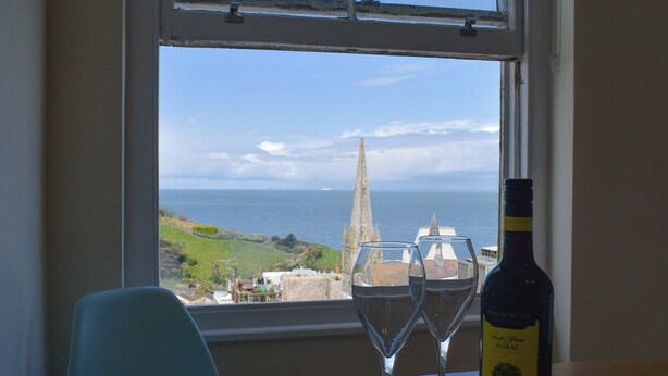 Una mesa con dos copas de vino y una botella, ubicada junto a una ventana con vistas a la costa y una aguja de iglesia.
