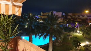 A nighttime view of a residential area with a swimming pool illuminated by blue lights, surrounded by palm trees and a few buildings in the background.