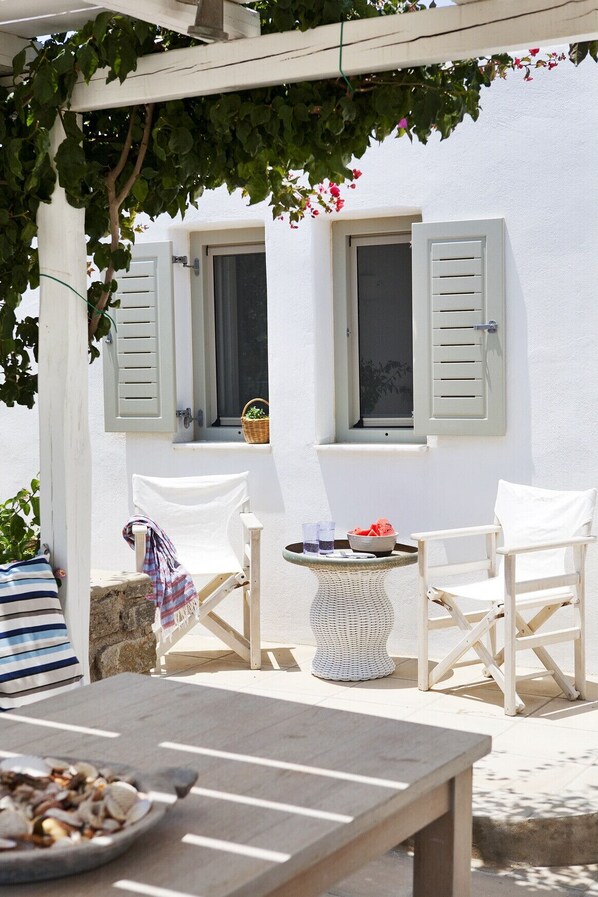 A patio with a table, two chairs, a wicker table, and a basket on a white wall with shutters.