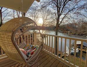 A wicker chair with a colorful cushion on a wooden deck overlooking a body of water with trees in the background. - Green Heron Cottage (Sherrills Ford)