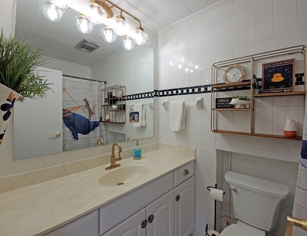 A modern bathroom with a white sink, gold faucet, and a mirror with lights above. There are shelves with toiletries and a clock.