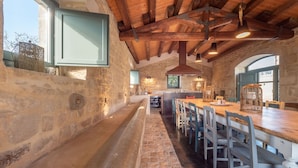 Une cuisine rustique aux murs de pierre, avec des poutres en bois et une longue table en bois entourée de chaises.