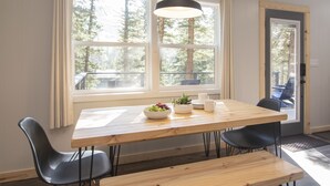 A dining area with a wooden table, black chairs, a hanging light fixture, and a view of trees through large windows.
