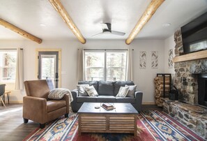 A cozy living room with a brown leather armchair, a gray sofa, and a wooden coffee table. There is a stone fireplace and a television mounted on the wall.