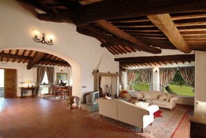 A spacious living room with a high ceiling, wooden beams, a white sofa, a dining area with a table and chairs, and large windows with curtains.