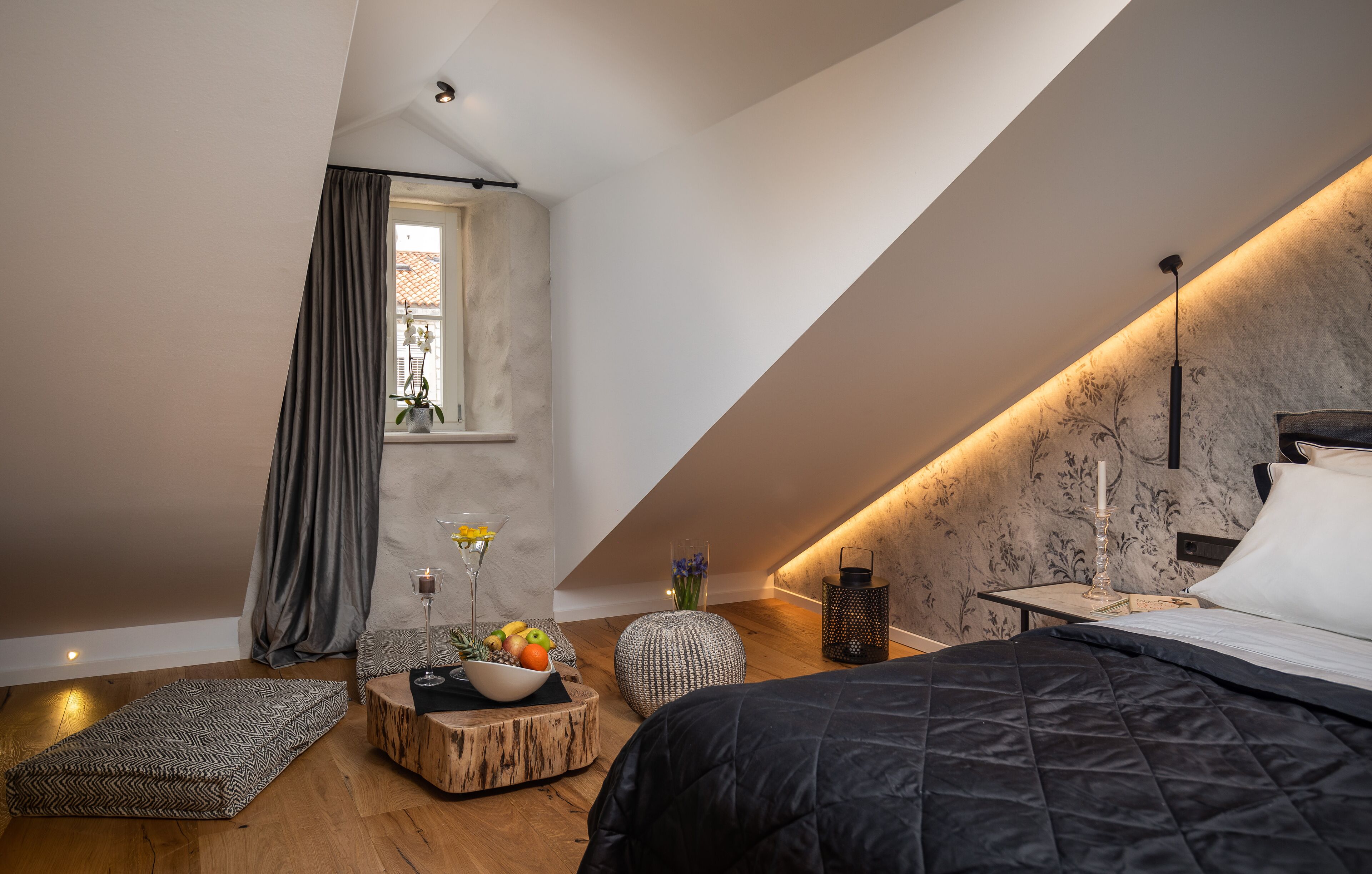 Een moderne slaapkamer met een schuine kap, een bed met een zwart sprei, een nachtkastje met een lamp, een klein rond tafeltje met fruit en bloemen, en een raam met gordijnen.