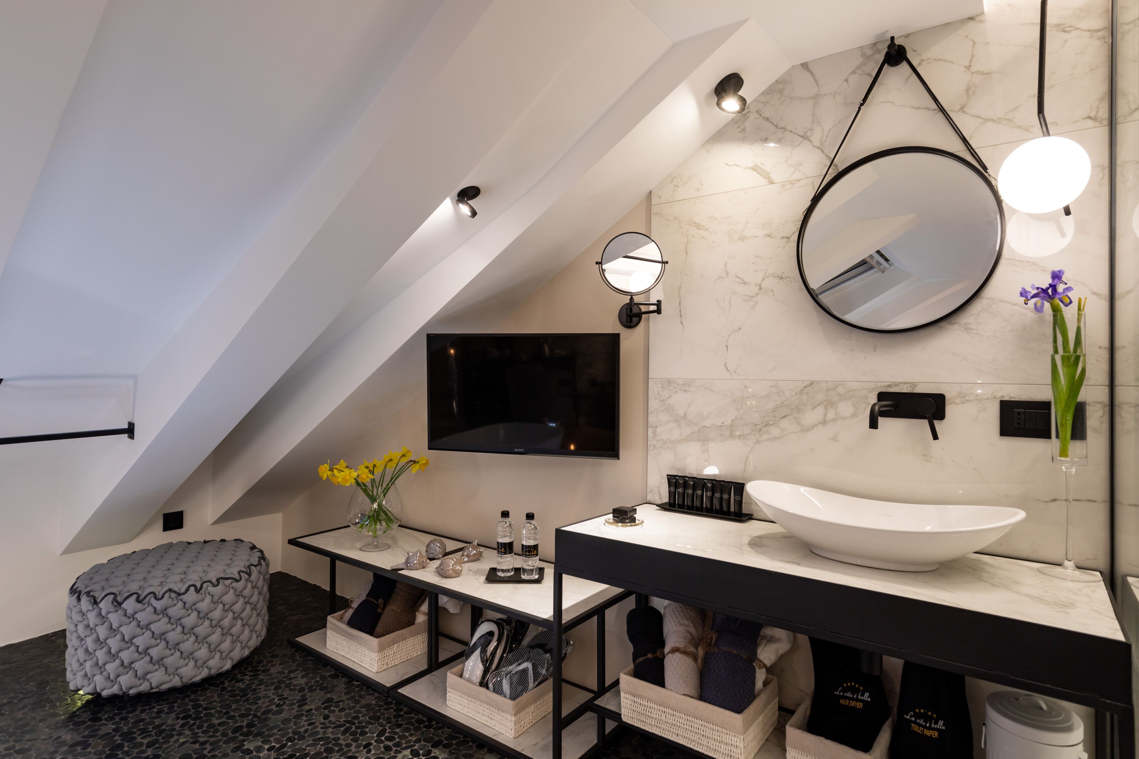 Een moderne badkamer met een grote witte wastafel, een ronde spiegel, een flatscreen televisie en een zithoekje met kussens.