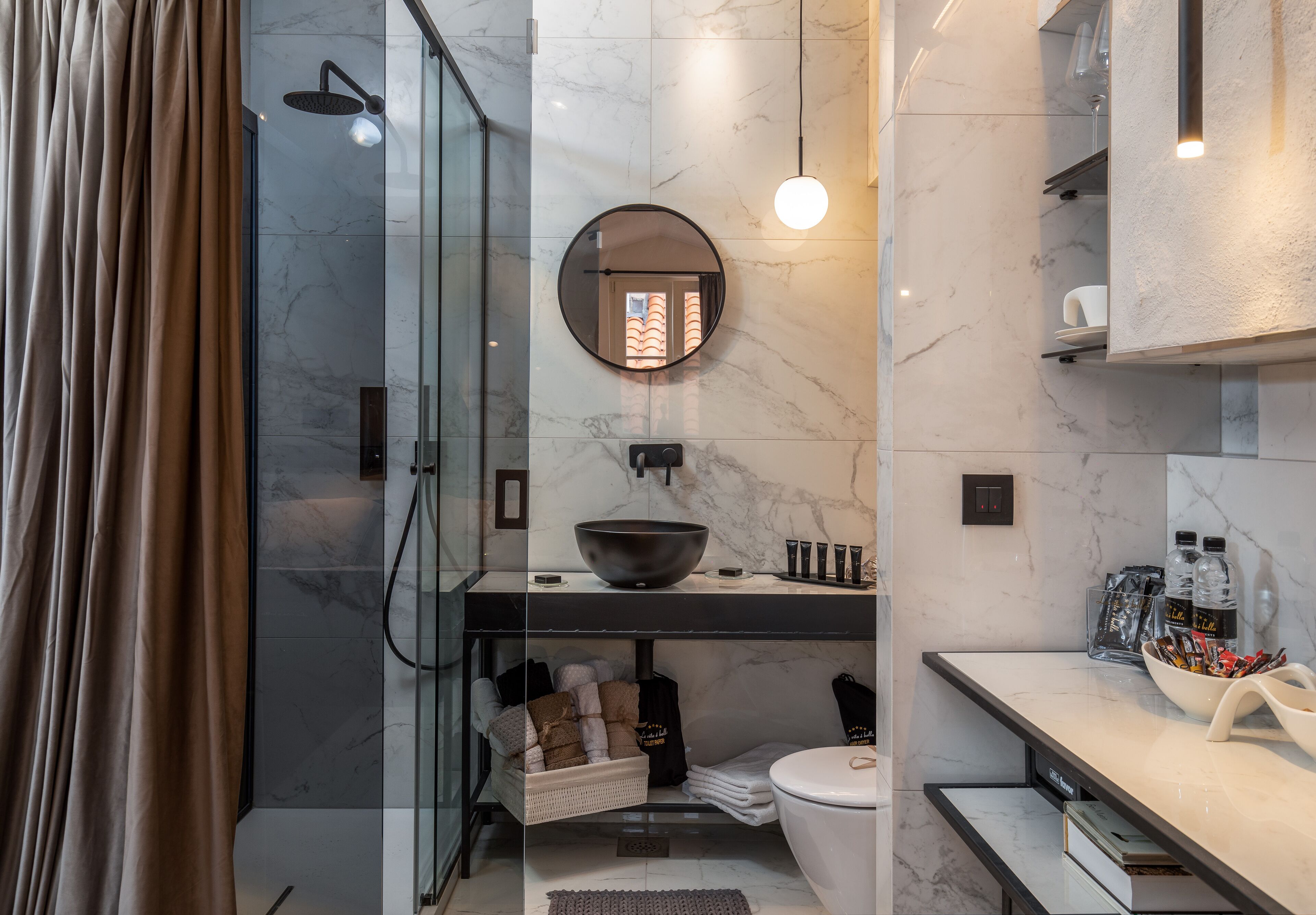 Une salle de bain moderne avec une douche à l’italienne en verre, un miroir rond, un lavabo noir en forme de cuve et un mur recouvert de carreaux de marbre.