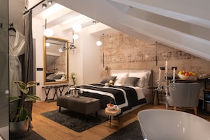 Quarto moderno com cama grande, área de estar com poltrona, banheira e mesa de jantar com frutas.