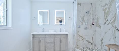 Un baño moderno con ducha de mármol, tocador con dos espejos y un pequeño banco de madera.