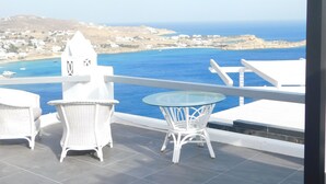 Ein Balkon mit weißen Korbmöbeln, einem Glastisch und Blick auf das Meer sowie entfernte Gebäude.