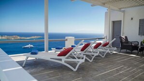 Une terrasse dotée de chaises longues blanches et de coussins rouges, offrant une vue sur une mer d’un bleu limpide et une île lointaine.