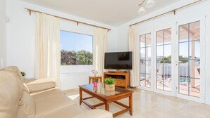 A living room with a beige sofa, a wooden coffee table with a plant, a flat-screen TV, and large windows with curtains.