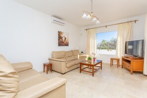 Ein Wohnzimmer mit einem beigen Sofa, einem Couchtisch, einem Fernseher auf einem Holzboden und einem Fenster mit Vorhängen.