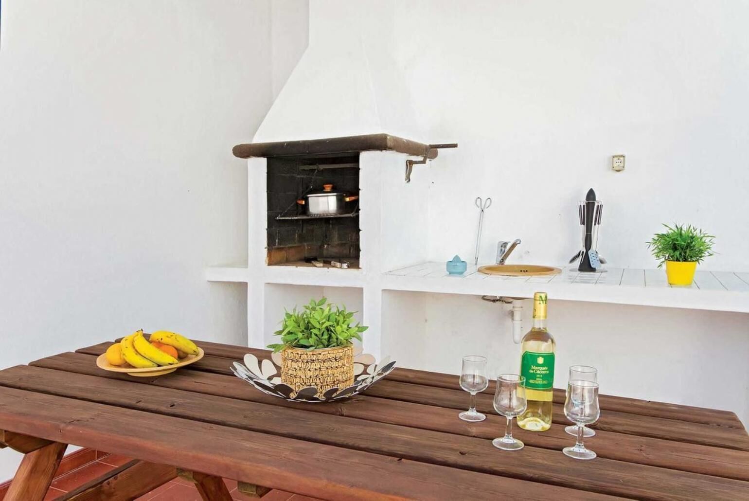 Ein Holztisch mit einer Bananenschale, einer Weinflasche und zwei Gläsern, einem gewebten Korb mit einer Pflanze und einem kleinen Regal mit Küchenutensilien und einem Blumentopf.