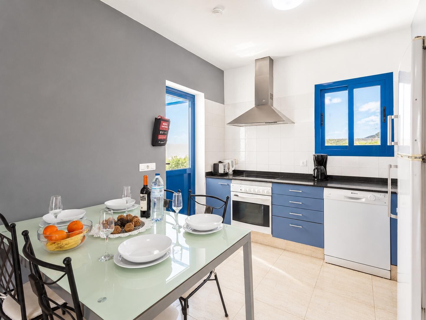 Een moderne keuken met blauwe kastjes, een roestvrijstalen afzuigkap en een eethoek met een tafel gedekt voor twee personen.