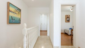 A hallway with a white railing, a painting on the wall, and a door leading to a room with a bed and a table.
