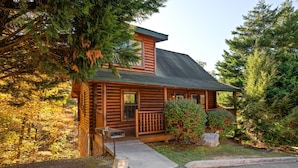 A log cabin with a porch, surrounded by trees and a small garden. - Cozy Cabin: Hot Tub, Pool Table, Jacuzzi, Sleeps 8 (Pigeon Forge)