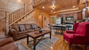 A wooden cabin interior with a staircase, a living room with a sofa and armchair, a kitchen with appliances, and a dining area. - Cozy Cabin: Hot Tub, Pool Table, Jacuzzi, Sleeps 8 (Pigeon Forge)