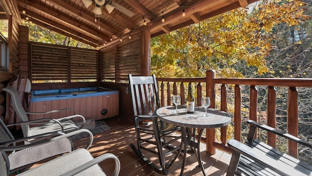 A wooden deck with a hot tub, rocking chairs, and a table with wine glasses. - Cozy Cabin: Hot Tub, Pool Table, Jacuzzi, Sleeps 8 (Pigeon Forge)