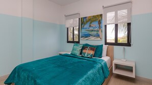 Een slaapkamer met een blauwgroene sprei, een nachtkastje, een wandtelevisie en een schilderij van een strandgezicht.