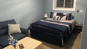 A bedroom with a bed, a nightstand, a bedside table, a lamp, a window with blinds, a wall-mounted air conditioner, and a small table with a vase and a book.