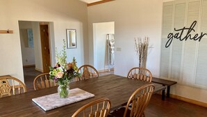 A dining room with a wooden table, six wooden chairs, a vase with flowers, and a 'gather' sign on the wall. - Spacious Farm House with Stunning Mountain Views (Joseph)