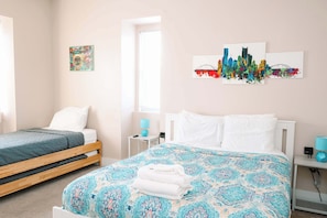 A bedroom with two beds, a window, artwork on the wall, and a bedside table with a lamp.
