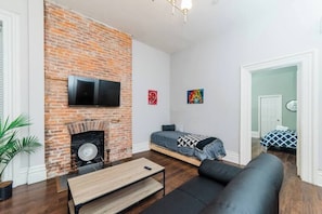 Una habitación con chimenea de ladrillo, una cama con colcha azul y blanca, un sofá negro y una mesa de centro de madera.