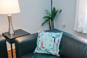 Una sala de estar moderna con un sofá de cuero negro, un cojín estampado, una lámpara sobre una mesita auxiliar y una planta en maceta.