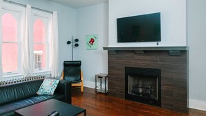Uma sala de estar com lareira, televisão montada acima dela, um sofá, uma poltrona e uma mesinha.
