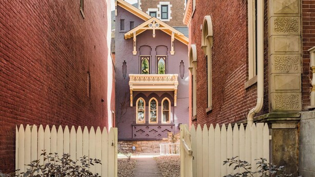 Uma viela estreita com cerca branca de piquete, um edifício roxo com janelas em arco e um muro de tijolos.